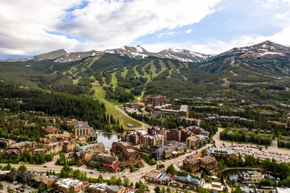 An aerial view of the town of Breckenridge, Colorado, in the foreground with lots of businesses, houses, and a ski resort, with green treed mountains in the backround and taller snow-capped mountains in the distance.